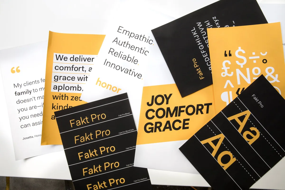 Gedrukte Honor-merkposters op een tafel: het woordmerk honor, lettertypespecimens in Fakt Pro, een citaatkaart en de woorden joy, comfort, grace in zwart en amber